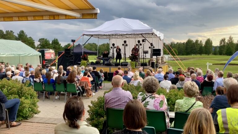Kesäinen ulkoilmakonsertti on golf-kentän reunalla. Yleisö istuu paikoillaan ja esiintyjät esiintyvät korkeammalla lavalla. Paikalla on ainakin sata henkilöä. Sää on pilvinen.