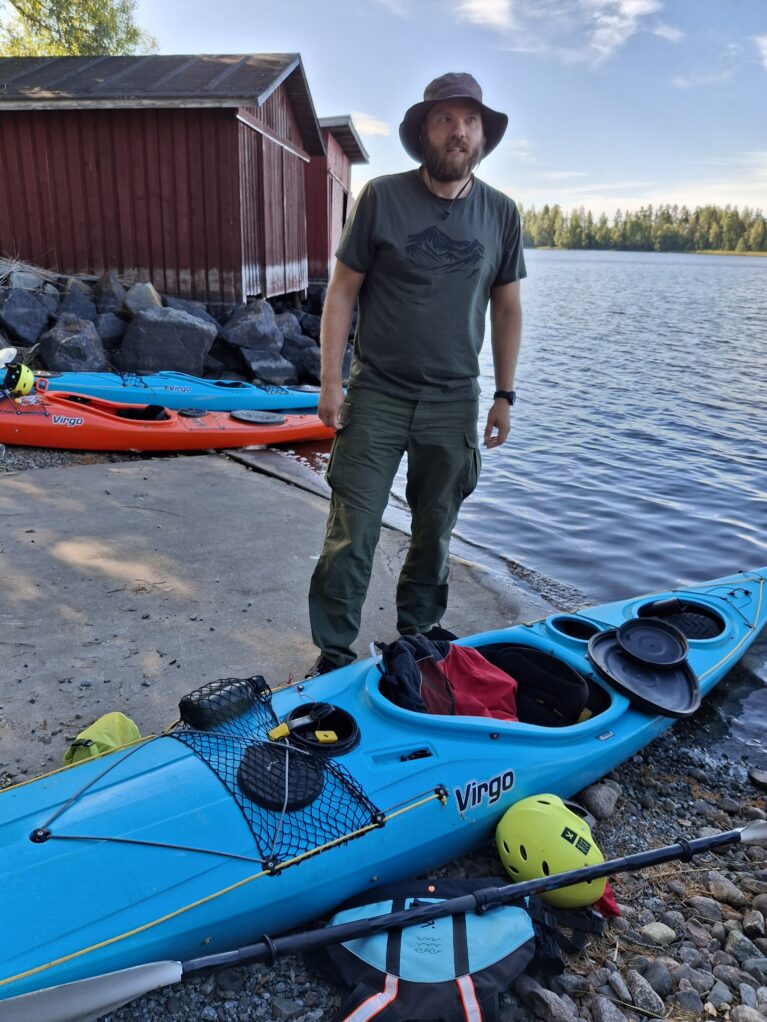 Mehtämikko valmistautui melomaan Paron sahanrannassa. Kuvassa lierihattuun ja ulkoiluvaatteisiin pukeutunut Mehtämikko ja sininen kajakki, takana rannan venevajoja ja sinistä järveä.