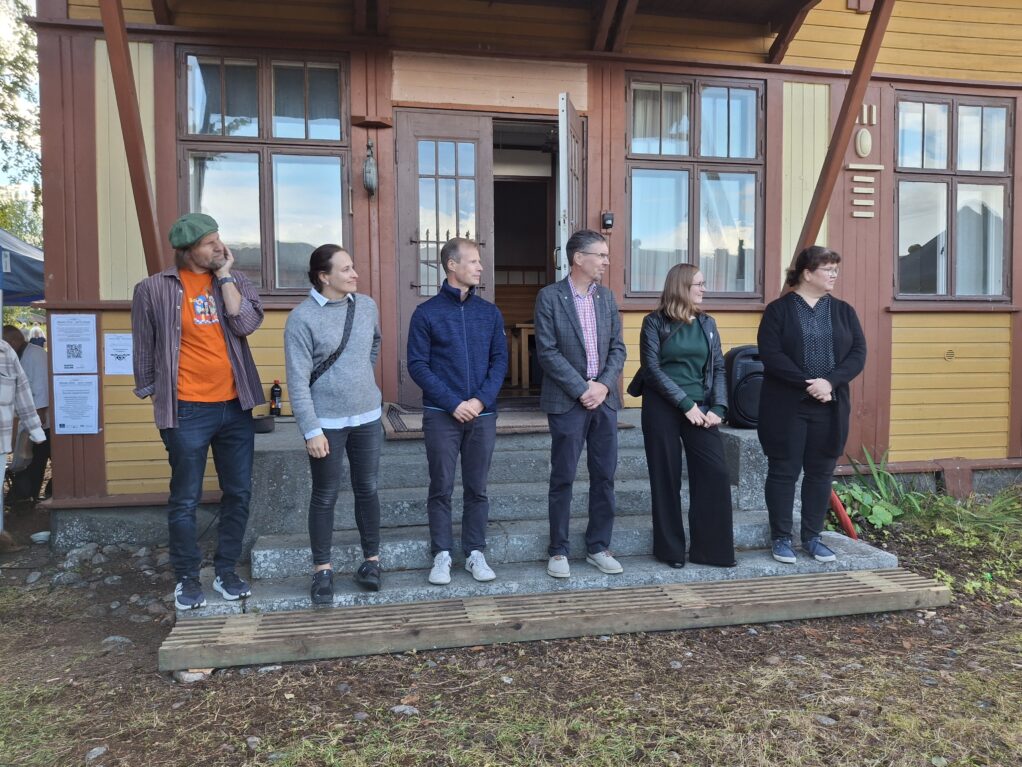 Kuusi vaikuttajaa seisoo rivissä Rantasalmen puisen, sinapinkeltaisen rautatieasemarakennuksen edessä kiviportailla.