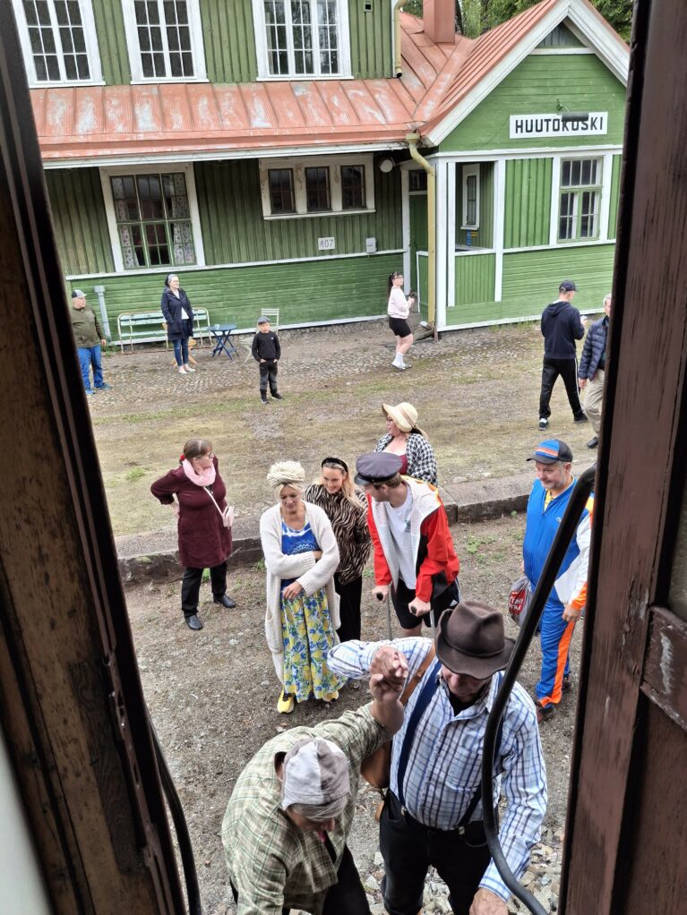 Huutokosken isokokoinen, vihreä puinen rautatieasemarakennus on taustalla, etualalla ihmisiä nousee höyryjunan kyytiin ja katselee junaa. Osa ihmisistä on pukeutunut historialliseen tyyliin.