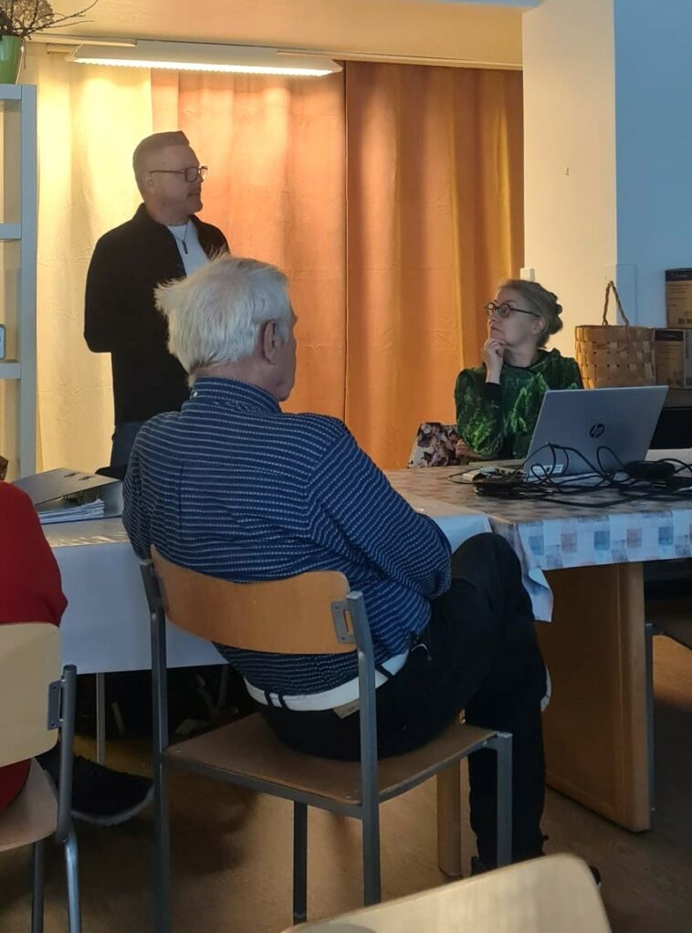 Yrittäjä Vesa Kärkkäinen seisoo taustalla, edessä pöydän ääressä istuvat Risto Salko ja projektikoordinaattori Anna Kaasinen. Huone on illan hämärä.